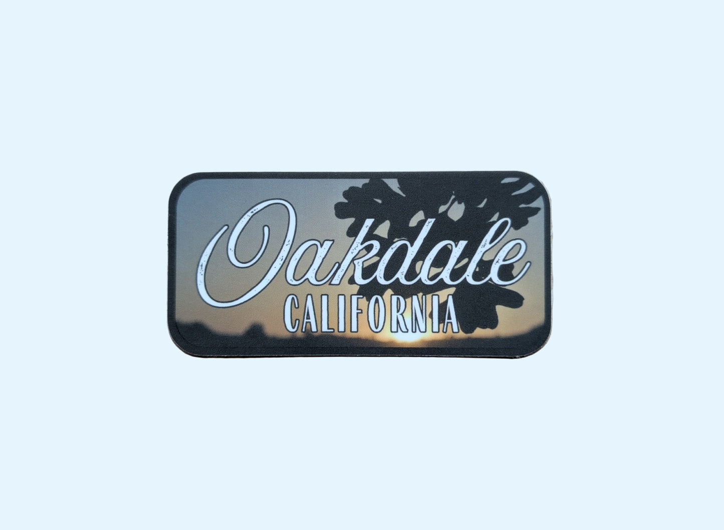 Rectangular sticker with dark gray border with photo background of oak leaves in front of sunset with the words "Oakdale California"
