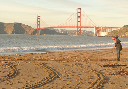 View of the Golden Gate Bridge from Baker Beach at sunset with woman raking sand in foreground