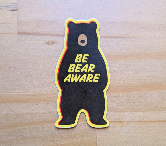 Yellow-bordered bear-shaped sticker that reads: Be Bear Aware with wood background