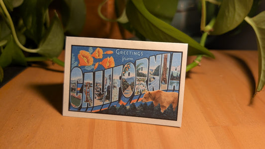 Mini California card with vintage postcard design on confetti white paper standing up on desk with plant in background