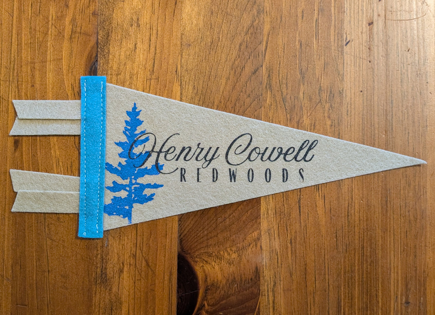 Mini khaki pennant with the words "Henry Cowell Redwoods" in black and a redwood in blue.