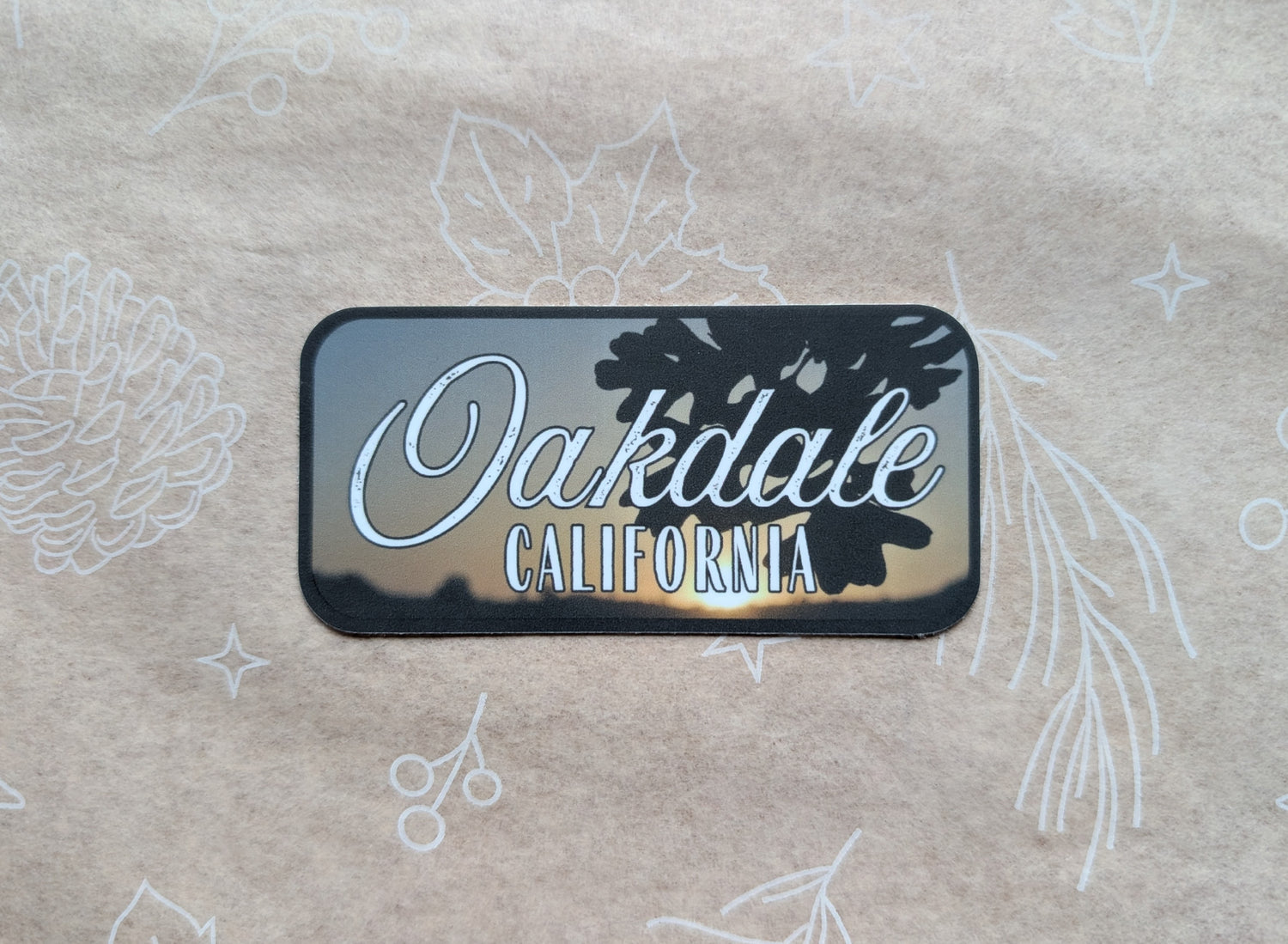 Rectangular sticker with dark gray border with photo background of oak leaves in front of sunset with the words "Oakdale California"